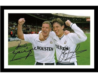 May include: A framed photograph of two football players in white jerseys with the text HOLSTEN and Hummel logos, celebrating on a green pitch with their arms raised. The image features two distinct black ink signatures across the players' chests.