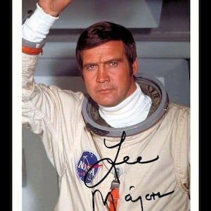 May include: A colour photograph of an astronaut in a white spacesuit with a NASA patch on the left side of the chest. The astronaut is holding onto a blue handrail. The astronaut is looking at the camera with a serious expression. The astronaut's signature, "Lee Majors", is written in black ink on the right side of the image.
