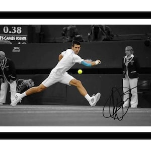 May include: A black and white photograph of a tennis player in a white shirt and shorts lunging to hit a bright yellow tennis ball. The player wears a blue arm sleeve and white trainers. A black signature is visible in the lower right corner.