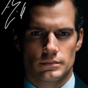 May include: A portrait of a man with dark hair and a stern expression. He is wearing a suit and a light blue collared shirt. The image includes a white signature in the upper left corner.
