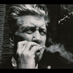 May include: Black and white photograph of a man smoking a cigarette. The man has a serious expression on his face and is wearing a dark suit. The photograph is signed "David Lynch".