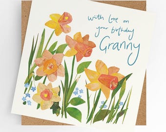Op de afbeelding: Een witte kaart met een bloemmotief met gele en oranje narcissen en blauwe vergeet-me-nietjes. De kaart heeft de tekst "with love on your birthday Granny" in blauwe inkt.