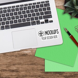 Może przedstawiać: Srebrny laptop z zielonym i jasnozielonym notesem i długopisem na drewnianej powierzchni. Laptop ma naklejkę z napisem "The Hobby Girl Mockups Your Design Here".
