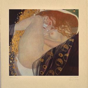 以下が含まれることがあります： グスタフ・クリムトの絵画「接吻」のプリント。長い赤い髪の女性が金と黒のベッドに横たわっている。女性は裸で、体は金箔で覆われている。絵画は右下に「グスタフ・クリムト」と署名されている。プリントは左下に「108/150」と番号が付けられている。