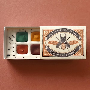 May include: A small rectangular box containing four square paint pans in green, yellow, red, and dark brown. The white tray has black speckles. The sliding cover features a beetle illustration and the text Earth Pigment Paints Lilian May Studio.