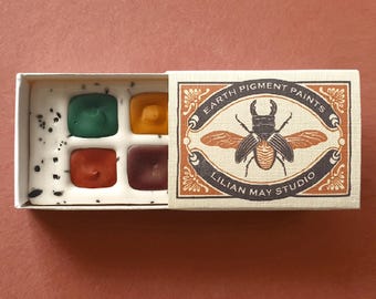 May include: A small rectangular box containing four square paint pans in green, yellow, red, and dark brown. The white tray has black speckles. The sliding cover features a beetle illustration and the text Earth Pigment Paints Lilian May Studio.