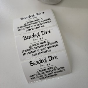 May include: Three white sticker labels with black text that reads "Beaded Item Care Tips" and a warning symbol. The text reads "Choking Hazard. Do not leave unattended with children. Not a teething toy. Do not put in mouth. Clean with damp rag. Lilmawnster's Designs."