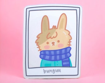 Peut inclure: Un autocollant représentant un lapin de dessin animé portant une écharpe rayée bleu et blanche. Le lapin est brun et blanc avec une chemise bleu clair. L'autocollant est dans un cadre blanc avec une bordure noire et le mot "bunguu" écrit en bas.