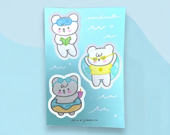 Peut inclure: Trois autocollants mignons de dessin animé d'ours sur un fond bleu. Les ours portent des lunettes de soleil et profitent d'activités estivales. Le texte "marshmallow" et "visitez-nous sur jrainbowsc.ca" figure également sur la feuille d'autocollants.