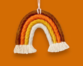 Peut inclure: Une suspension murale en macramé arc-en-ciel avec du fil brun, orange, jaune et blanc. La suspension a une frange en bas.