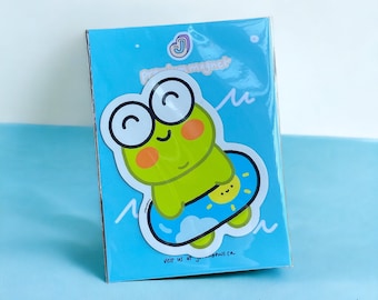 Peut inclure: Un personnage de dessin animé de grenouille verte portant une bouée gonflable bleue et jaune avec un soleil et des nuages. La grenouille sourit et a de grands yeux. L'arrière-plan est bleu avec des vagues blanches. Le texte "MAGNET!" est écrit en noir en bas de l'image.