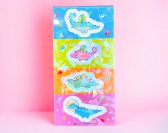 Peut inclure: Quatre autocollants holographiques colorés représentant des dinosaures portant des cadeaux. Les autocollants sont disposés verticalement sur un fond rose. Les dinosaures sont bleus, roses, verts et bleus. Le texte "let us be friends" est en bas des autocollants.