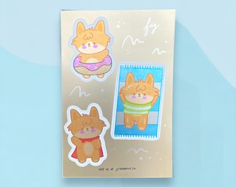 Peut inclure: Trois autocollants représentant un renard de dessin animé dans différentes activités estivales. Le premier autocollant montre le renard portant un beignet rose comme dispositif de flottaison. Le deuxième autocollant montre le renard allongé sur une serviette rayée. Le troisième autocollant montre le renard portant une cape rouge. Les autocollants sont sur un fond doré avec le texte "visit us at jsrainbows.ca".