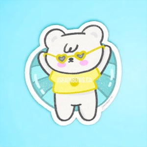 May include: A white bear wearing heart-shaped sunglasses and a yellow shirt with the text "jsrainbows.ca" is floating on a blue and white inflatable ring.