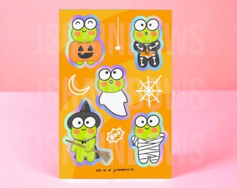 Peut inclure: Une feuille d'autocollants en vinyle imperméable représentant une grenouille verte vêtue de différents costumes d'Halloween. La grenouille est habillée en citrouille, en fantôme, en sorcière, en squelette et en momie. Les autocollants sont sur un fond orange avec le texte "Autocollant en vinyle imperméable" en bas.