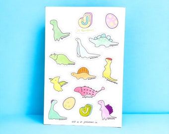 Peut inclure: Une feuille d'autocollants de dinosaures colorés avec le texte "JS Rainbows" et "visitez-nous sur jsrainbows.ca". Les autocollants présentent différents dinosaures, dont un stégosaure, un tricératops, un brachiosaure et un ankylosaure.