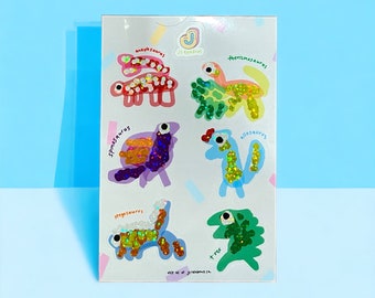 Peut inclure: Une feuille d'autocollants de dinosaures colorés avec des accents de paillettes. Les autocollants présentent les noms de chaque dinosaure : Ankylosaurus, Therizinosaurus, Spinosaurus, Allosaurus, Stegosaurus et T-Rex.