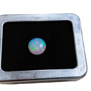 Pode incluir: Uma pedra preciosa de opala redonda e iridescente, exibida numa pequena caixa de metal retangular com interior de veludo preto. A opala tem uma mistura de cores, incluindo azul, verde e rosa.