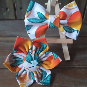 May include: Two fabric hair accessories: a bow and a flower. The bow is orange, yellow, and green with a white background. The flower is orange, yellow, and green with a white background and a white flower center. Both are on a wooden surface.