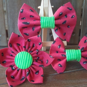 May include: Three handmade fabric accessories: two bow ties and a flower. The bow ties and flower are red with black seed details, and green striped centers. The accessories are likely for pets or hair.