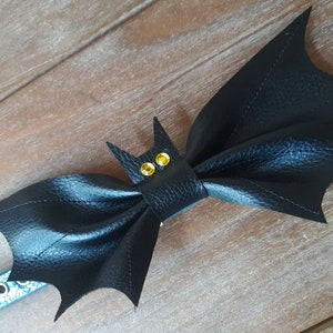 May include: A black bat-shaped pet collar accessory with yellow gem eyes. The bat wings are textured and stitched. The collar is blue with white snowflake designs and silver eyelets. The accessory is on a wooden surface.