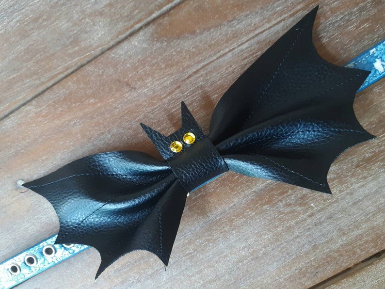 May include: A black bat-shaped pet collar accessory with yellow gem eyes. The bat wings are textured and stitched. The collar is blue with white snowflake designs and silver eyelets. The accessory is on a wooden surface.