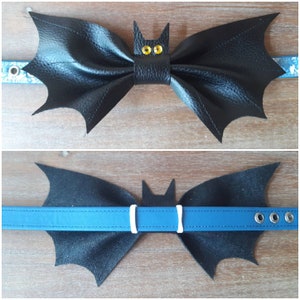May include: Black leather bat-shaped bow tie with yellow eye accents, attached to a blue and patterned collar. The bat has textured wings and a small bat head in the center. The collar has metal eyelets.
