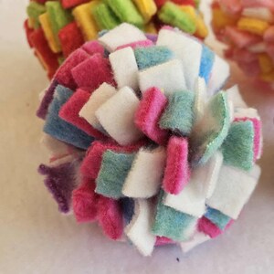 May include: Close-up of a colorful, handmade felt ball. The ball is composed of small, square felt pieces in various colors, including pink, white, blue, and green. Other felt balls are visible in the background, with similar designs and colors.