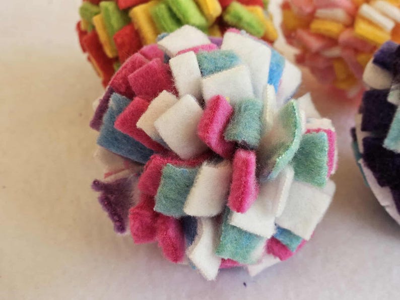 May include: Close-up of a colorful, handmade felt ball. The ball is composed of small, square felt pieces in various colors, including pink, white, blue, and green. Other felt balls are visible in the background, with similar designs and colors.