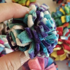 May include: A close-up of a handmade, colorful pom-pom. The pom-pom is held in a hand and is made of felt strips in shades of teal, purple, and white. Other pom-poms in various colors are visible in the background.