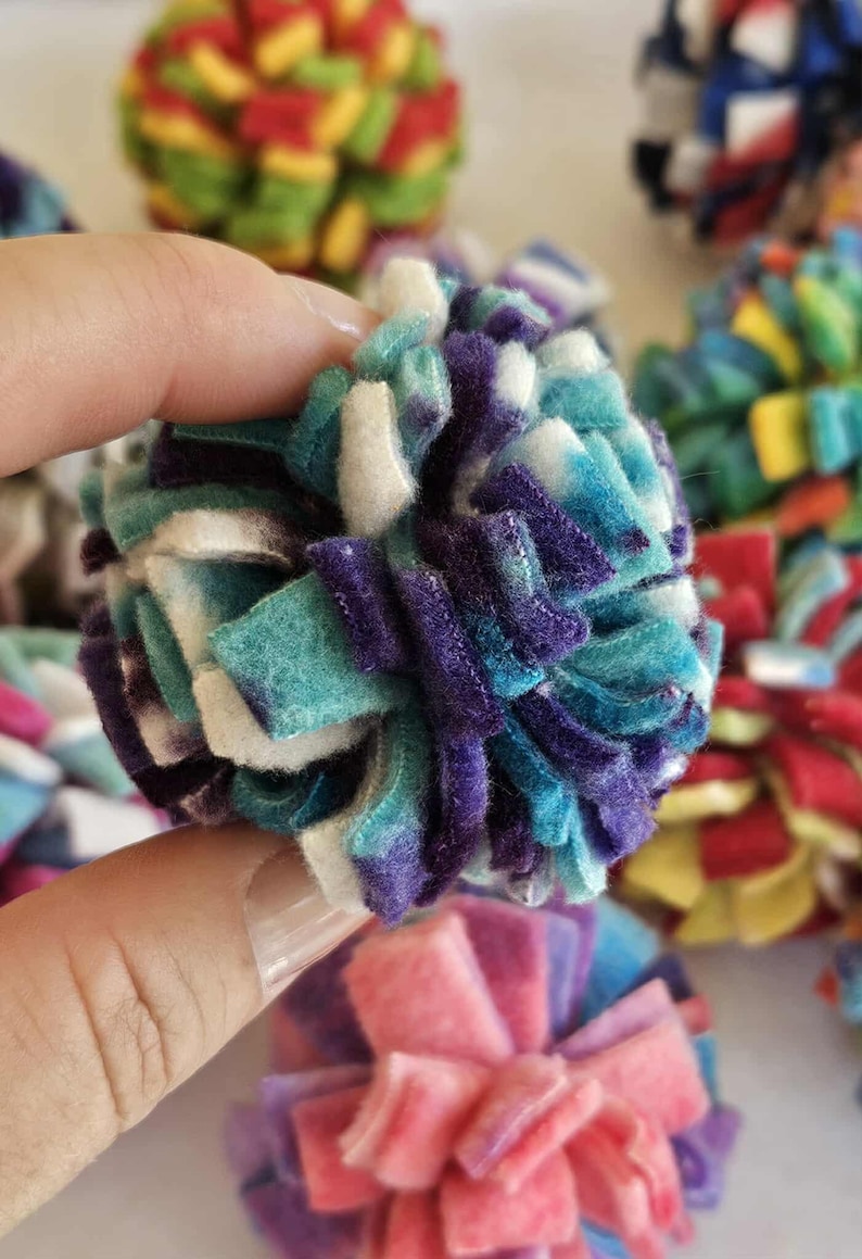 May include: A close-up of a handmade, colorful pom-pom. The pom-pom is held in a hand and is made of felt strips in shades of teal, purple, and white. Other pom-poms in various colors are visible in the background.