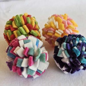 May include: Four colorful felt ball toys. One ball is red, yellow, and green. Another is pink, yellow, and white. The third is white, pink, and teal. The last is blue, purple, and white. These are likely cat toys.