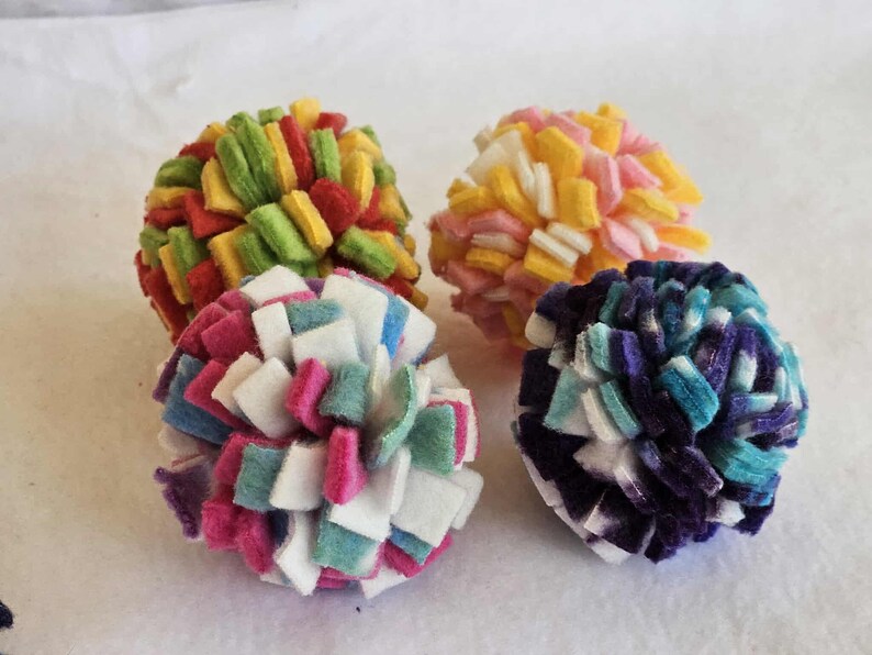 May include: Four colorful felt ball toys. One ball is red, yellow, and green. Another is pink, yellow, and white. The third is white, pink, and teal. The last is blue, purple, and white. These are likely cat toys.
