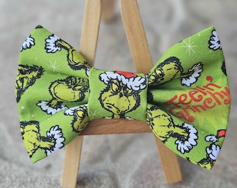May include: A green bow tie with a Grinch character design wearing a Santa hat and white snowflakes. The bow tie has the text "Feelin' Grinchy" in red. The bow tie is displayed on a small wooden easel.