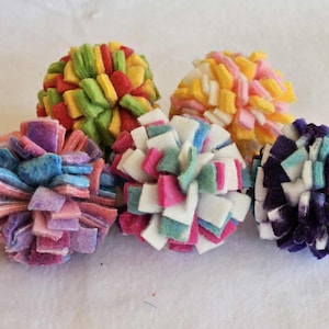 May include: Six colorful felt pom-poms in various colors, including red, green, yellow, pink, blue, and purple. Each pom-pom is made of small felt squares arranged in a spherical shape. The pom-poms are displayed on a white surface.