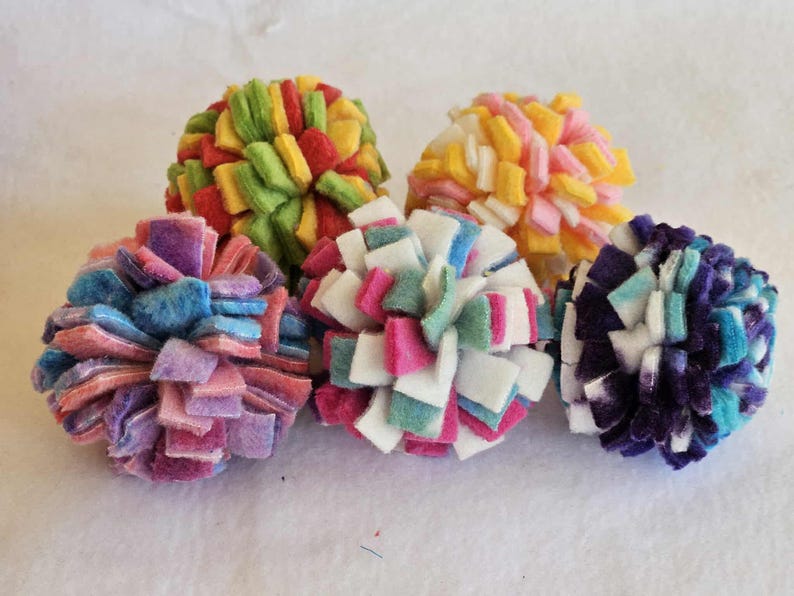 May include: Six colorful felt pom-poms in various colors, including red, green, yellow, pink, blue, and purple. Each pom-pom is made of small felt squares arranged in a spherical shape. The pom-poms are displayed on a white surface.