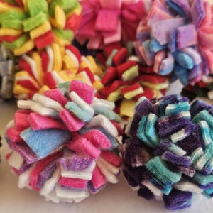 May include: A collection of colorful, handmade pom-poms in various sizes and hues. The pom-poms are made from soft, felt-like material and feature a layered, textured design. Colors include pink, blue, green, yellow, and purple.