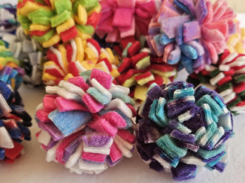 May include: A collection of colorful, handmade pom-poms in various sizes and hues. The pom-poms are made from soft, felt-like material and feature a layered, textured design. Colors include pink, blue, green, yellow, and purple.