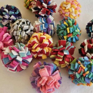 May include: A collection of colorful felt pom-poms in various sizes and hues, including pink, blue, red, yellow, and white. These handmade pom-poms are arranged on a white surface, showcasing their vibrant textures.