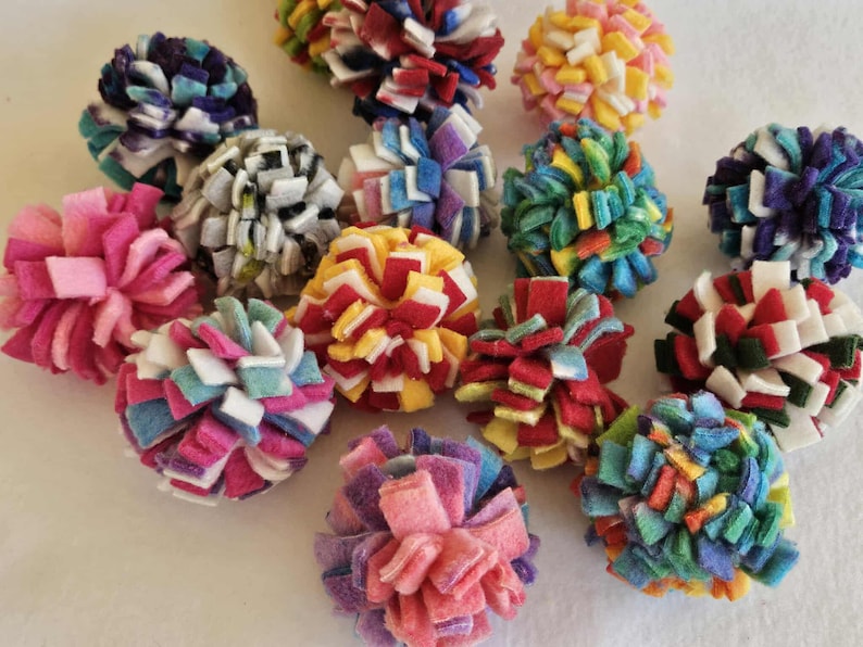 May include: A collection of colorful felt pom-poms in various sizes and hues, including pink, blue, red, yellow, and white. These handmade pom-poms are arranged on a white surface, showcasing their vibrant textures.