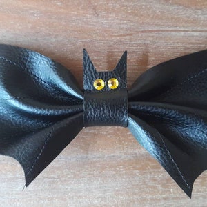 May include: A black leather bat-shaped pet collar accessory. The bat has outstretched wings and a central head with yellow gem eyes. The collar is blue with a white floral pattern and silver eyelets. A Halloween-themed pet accessory.