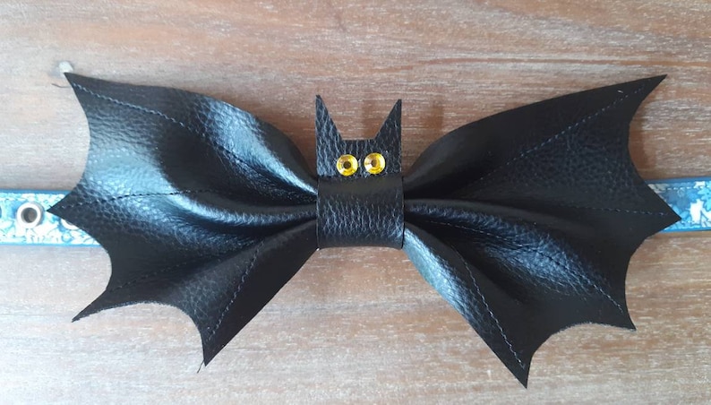 May include: A black leather bat-shaped pet collar accessory. The bat has outstretched wings and a central head with yellow gem eyes. The collar is blue with a white floral pattern and silver eyelets. A Halloween-themed pet accessory.