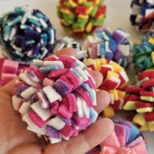 May include: Close-up of colorful, handmade felt balls in various colors, including pink, purple, white, blue, and yellow. The balls are made of small felt squares and are clustered together. The image shows several felt balls in focus.