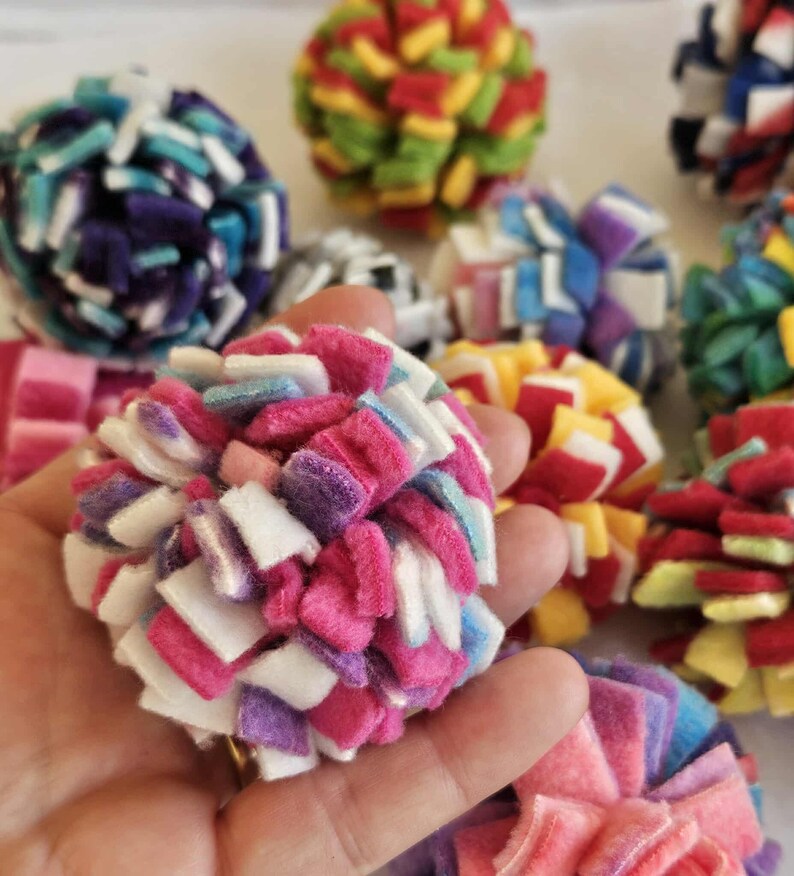 May include: Close-up of colorful, handmade felt balls in various colors, including pink, purple, white, blue, and yellow. The balls are made of small felt squares and are clustered together. The image shows several felt balls in focus.
