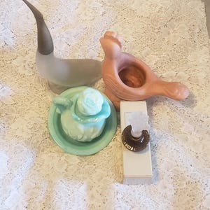 May include: A collection of vintage decorative items, including a frosted glass seal figurine, a peach-colored bird-shaped holder, a turquoise trinket dish with a rose, and a perfume bottle with a brown and clear stopper.
