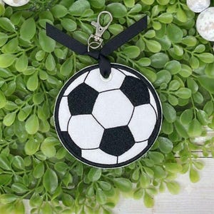 May include: A circular embroidered patch featuring a classic black and white soccer ball pattern. The patch is attached to a black ribbon loop with a silver-toned metal lobster claw clasp, allowing it to function as a decorative keychain or charm.