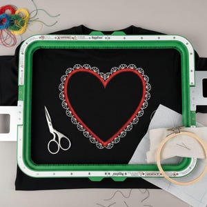 Może przedstawiać: Czarny t-shirt z czerwonym haftem w kształcie serca, oprawiony w zieloną tamborkę. Widoczne są również nożyczki, nici do haftowania oraz mniejsza drewniana tamborka z tkaniną, co sugeruje motyw rzemieślniczy.