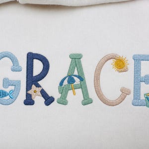 May include: The name GRACE is embroidered in colourful letters on a white fabric background. Each letter features beach-themed details: a blue fish on the G, a starfish on the R, a beach umbrella on the A, a sun on the C, and a bucket on the E.