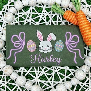May include: An olive green Easter-themed shirt with a white bunny, decorated eggs, and the name "Harley" embroidered in cursive. The design includes pink bows and is surrounded by decorative elements.