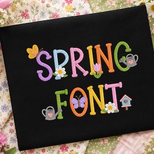 Könnte beinhalten: Eine schwarze Stoffoberfläche mit buntem Sticktext, der SPRING FONT lautet. Die Buchstaben sind mit kleinen Stickmotiven verziert, darunter ein gelber Schmetterling, weiße Gänseblümchen, eine rosa Tulpe, eine graue Gießkanne und ein blaues Vogelhaus.